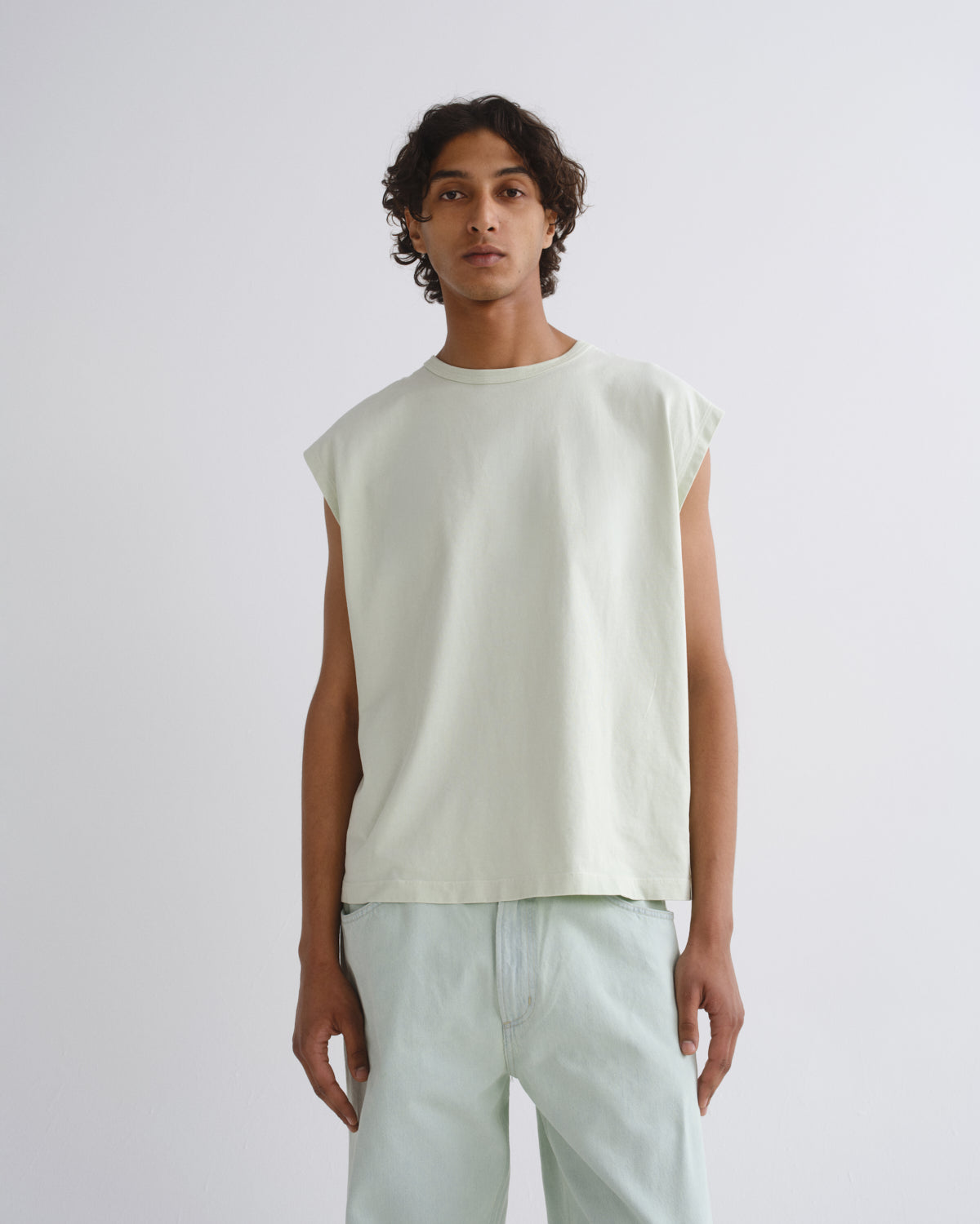 A man wearing a pale sage green sleeveless t-shirt and matching light sage green pants, standing against a light gray studio background.