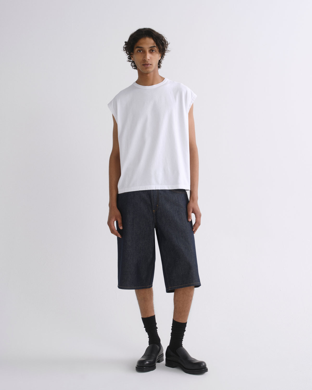 A model stands against a light gray studio background wearing a white sleeveless t-shirt, dark indigo denim shorts, black socks, and black leather boots.