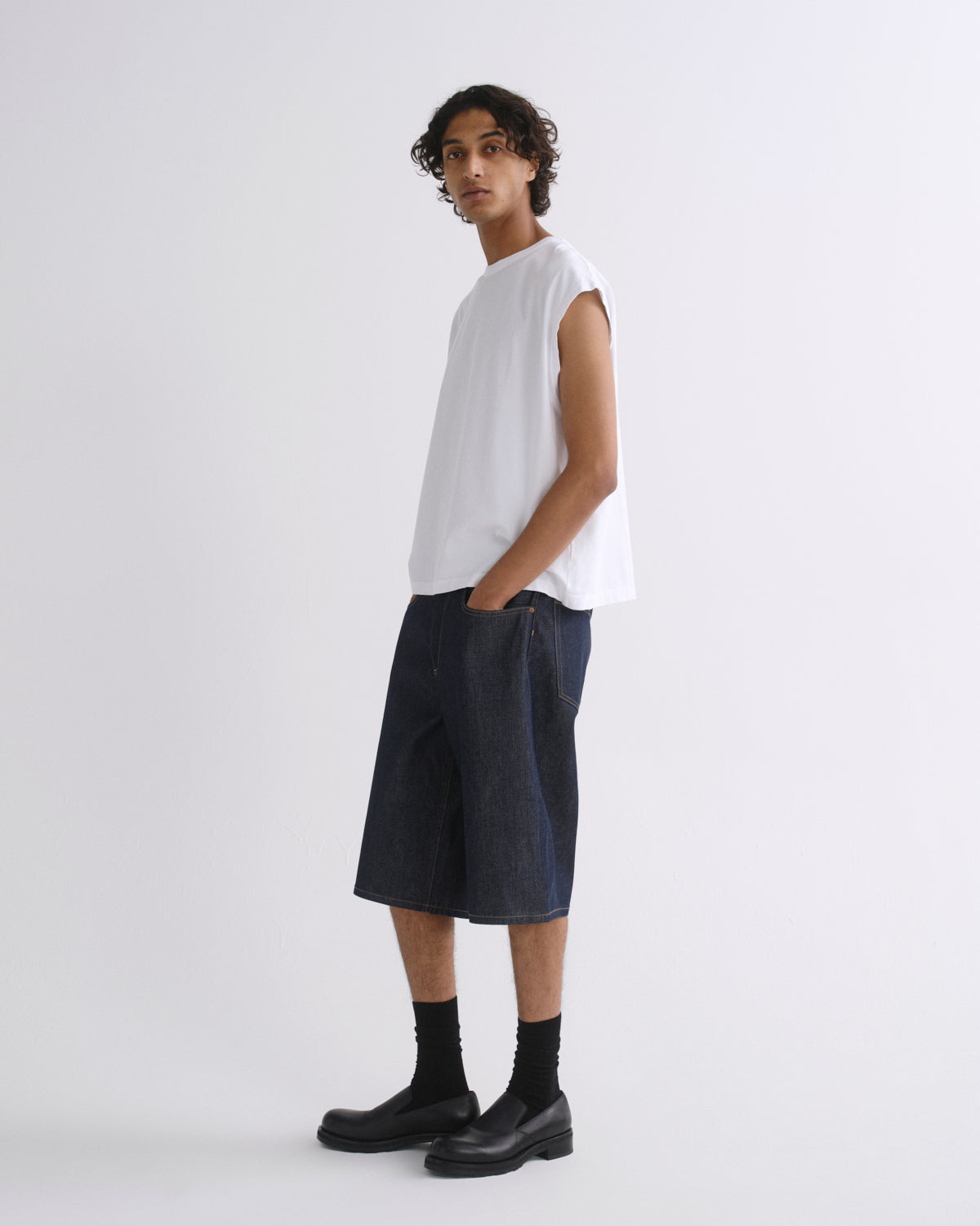 A model wearing a white sleeveless t-shirt, dark blue denim shorts, black socks, and black leather boots, standing against a plain light gray studio background.