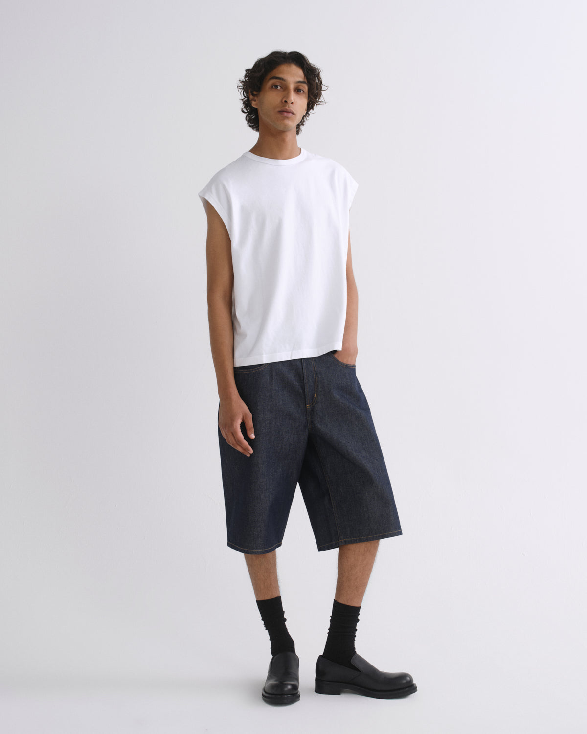 A model stands against a light gray studio background wearing a white sleeveless crewneck t-shirt, dark indigo denim shorts, black socks, and black leather boots.