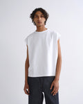 A man wearing a white sleeveless crewneck t-shirt and dark gray pants, standing against a light gray studio background.
