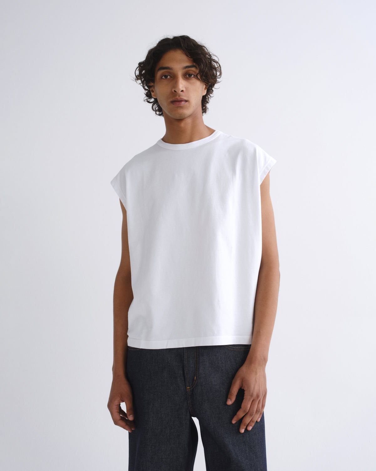 A man wearing a white sleeveless crewneck t-shirt and dark gray pants, standing against a light gray studio background.