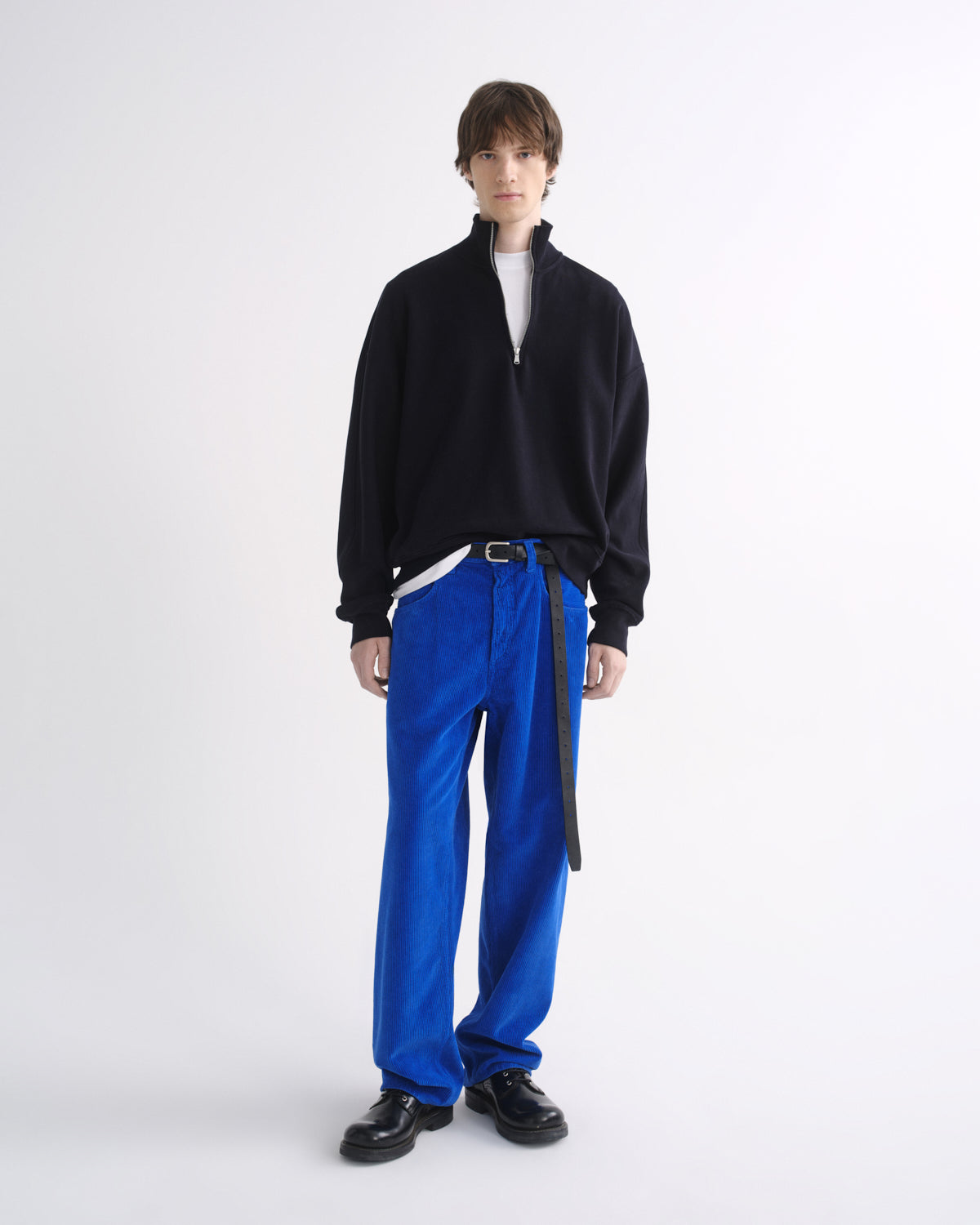 Person wearing a black zip-up jacket with bright blue pants and black shoes on a grey background