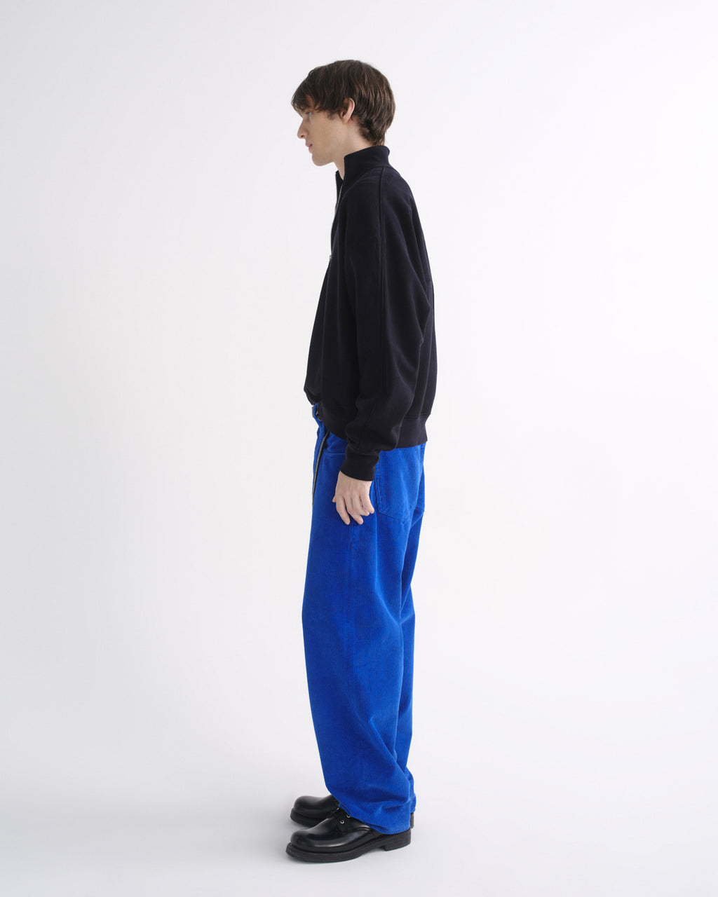 Person wearing a black zip-up jacket with bright blue pants and black shoes, side view, on a grey background