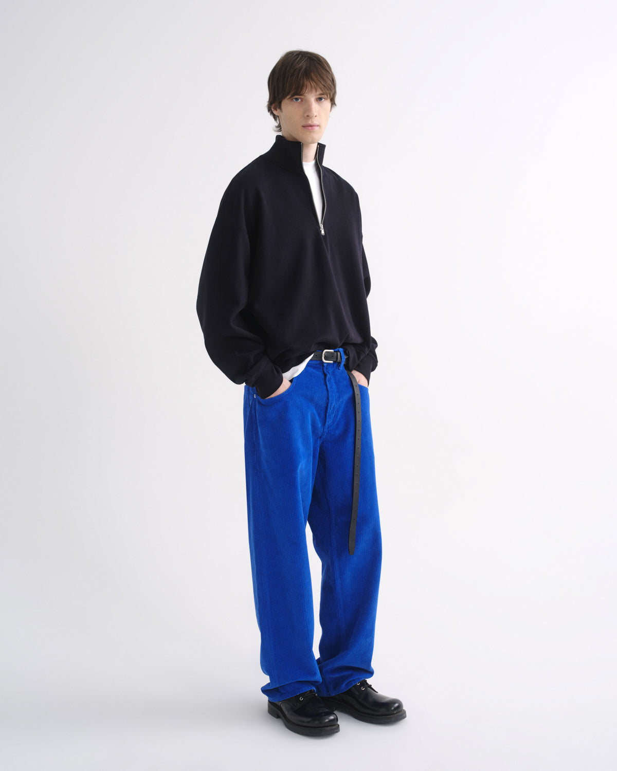 Person wearing a black zip-up jacket with bright blue pants and black shoes, side view, on a grey background