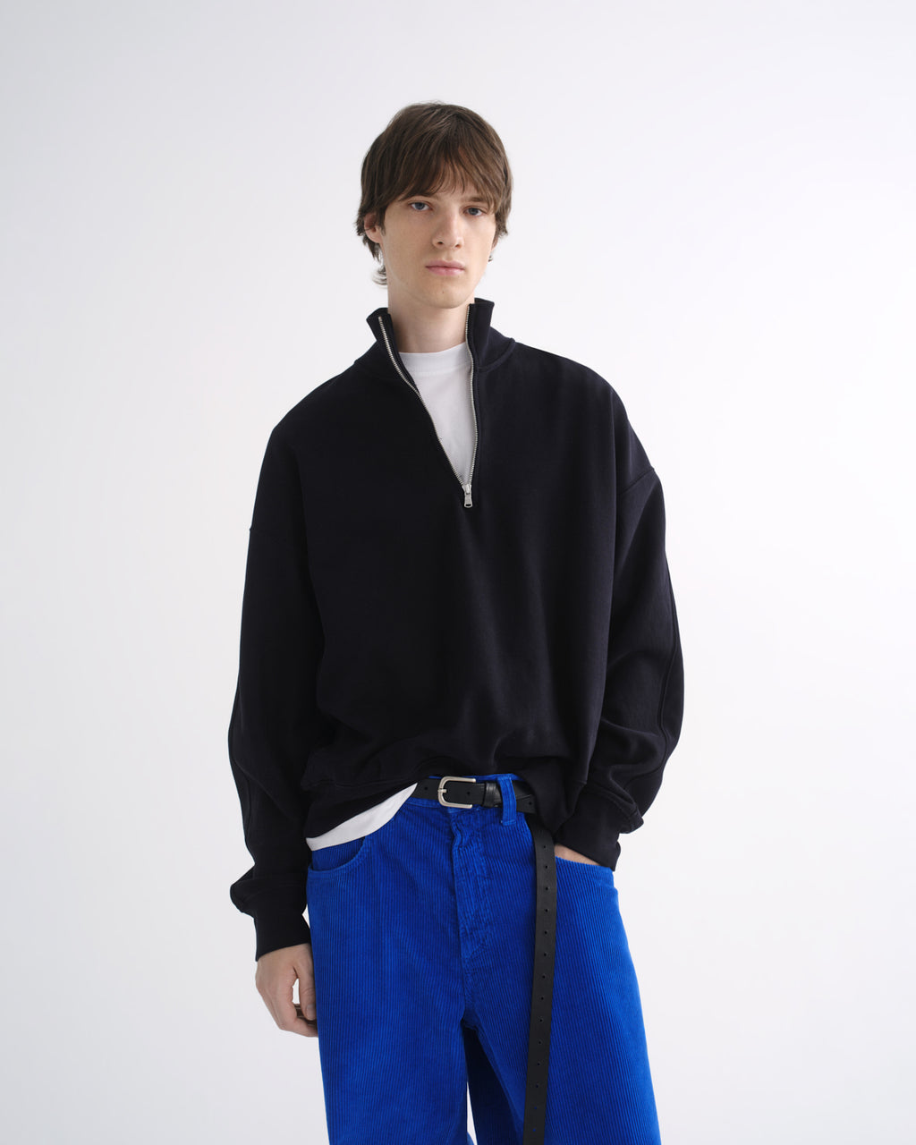 Person wearing a black zip-up jacket with bright blue pants and black shoes on a grey background