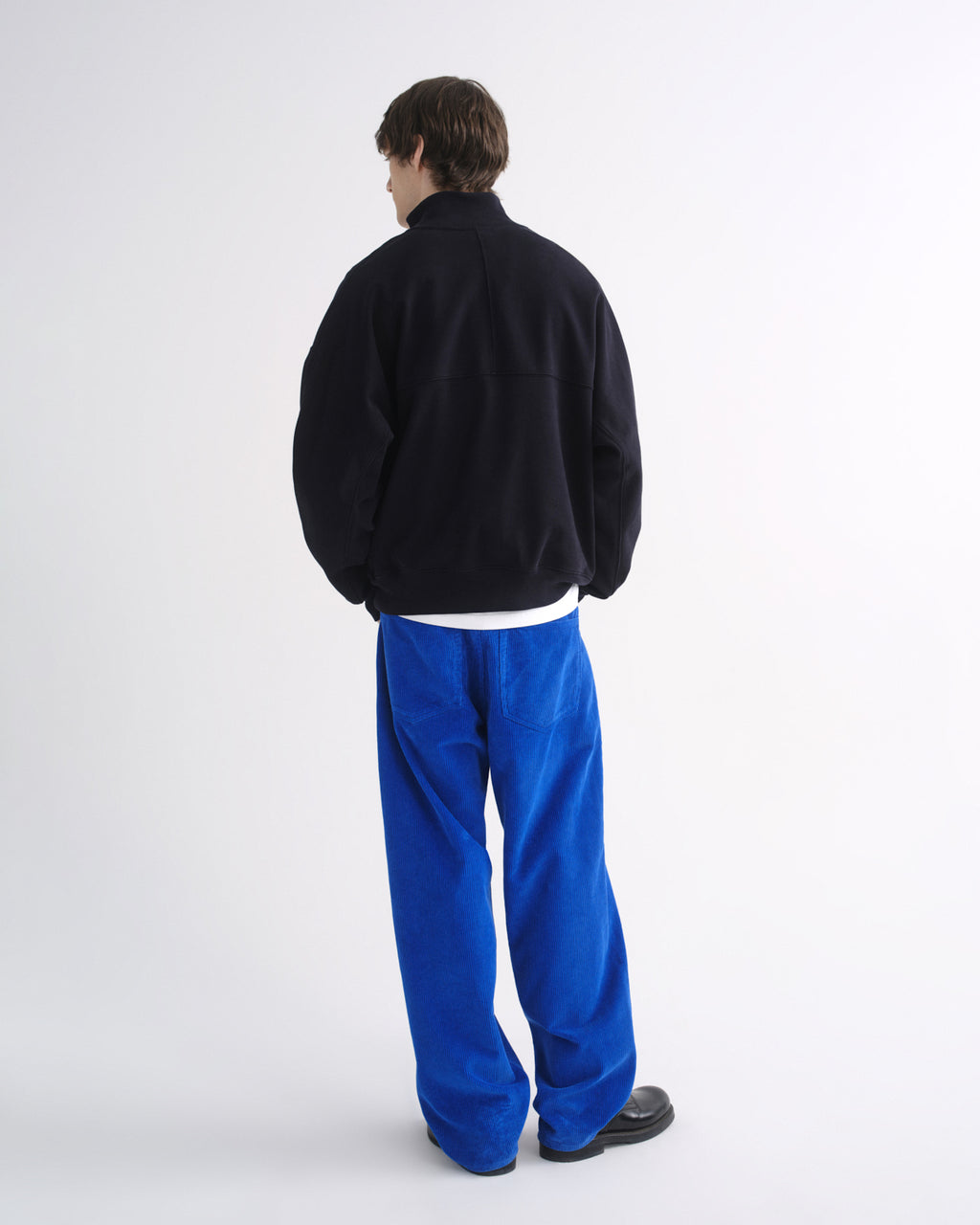 Person wearing a black zip-up jacket with bright blue pants and black shoes, back view, on a grey background