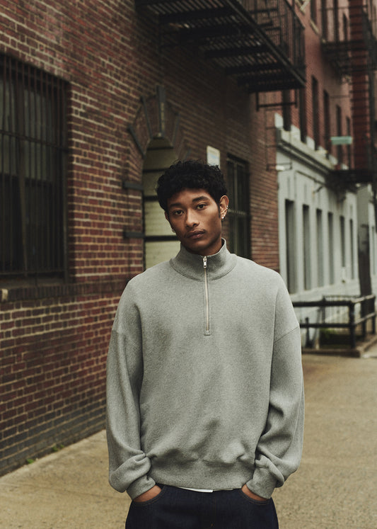 Man wearing a gray sweater standing against a brick wall.