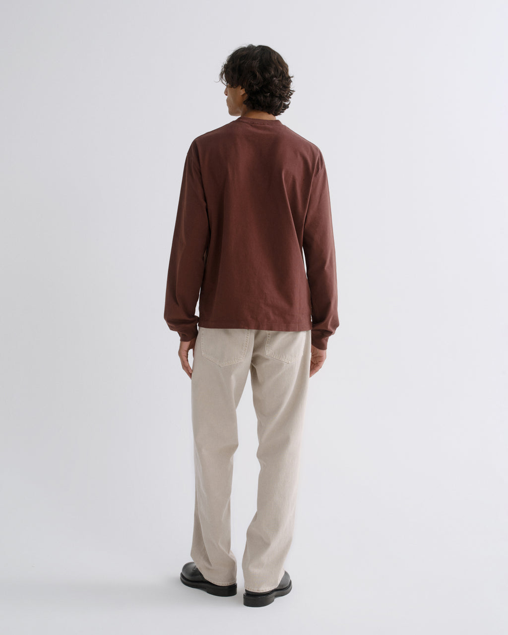 Man wearing a brown crewneck sweatshirt with cream straight leg pants viewed from the back on a grey background.