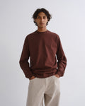 Man wearing a brown crewneck sweatshirt with cream straight leg pants on a grey background.