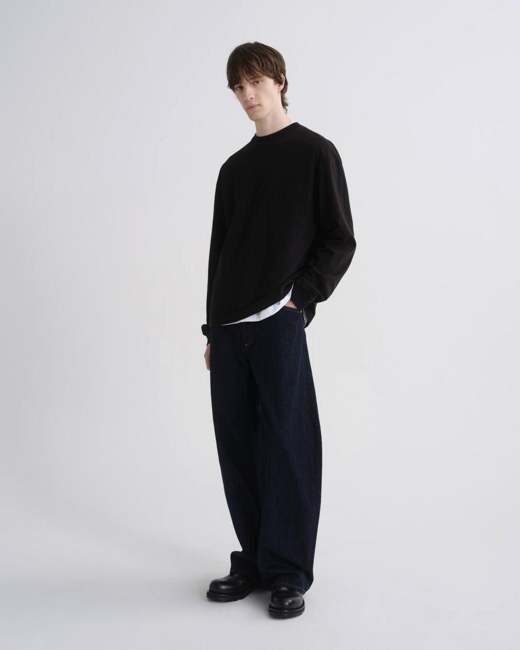 A model stands in a studio wearing a black long-sleeve crewneck sweater, dark navy wide-leg trousers, and black shoes, layered over a white undershirt.
