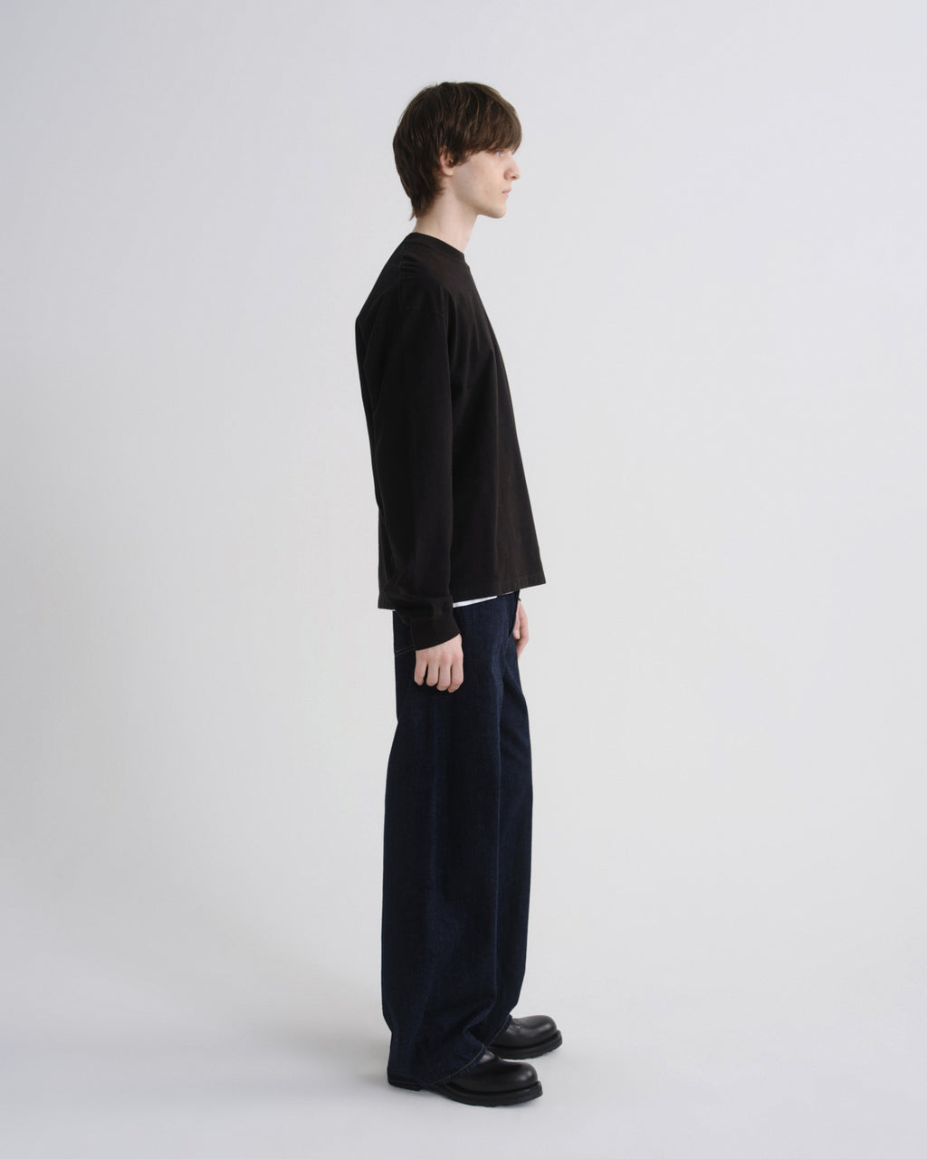 A model shown in profile wearing a black long-sleeve crewneck top, dark navy wide-leg trousers, and black shoes, standing against a light gray studio background.