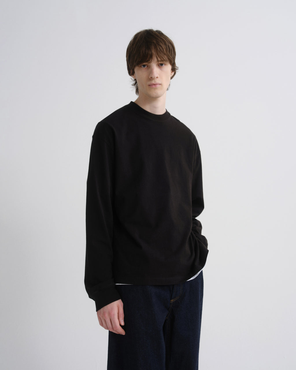 A young man with brown hair stands against a light gray studio background wearing a black long-sleeve crewneck sweatshirt and dark blue jeans.
