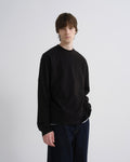 A young man with brown hair stands against a light gray studio background wearing a black long-sleeve crewneck sweatshirt and dark blue jeans.