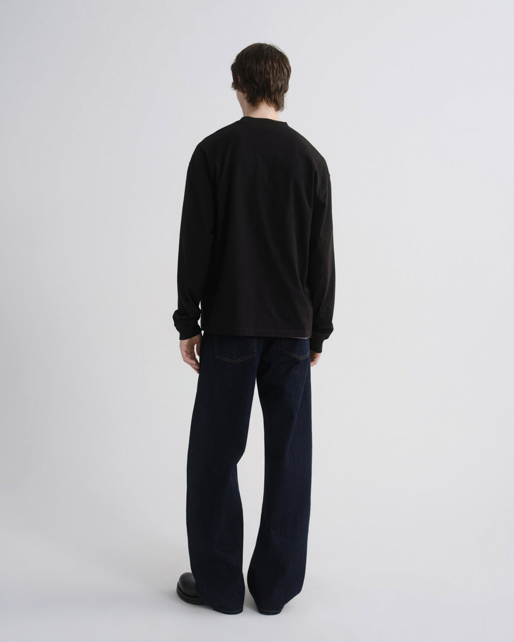 A model shown from the back wearing a black long-sleeve crewneck sweatshirt and dark navy wide-leg jeans against a light gray studio background.
