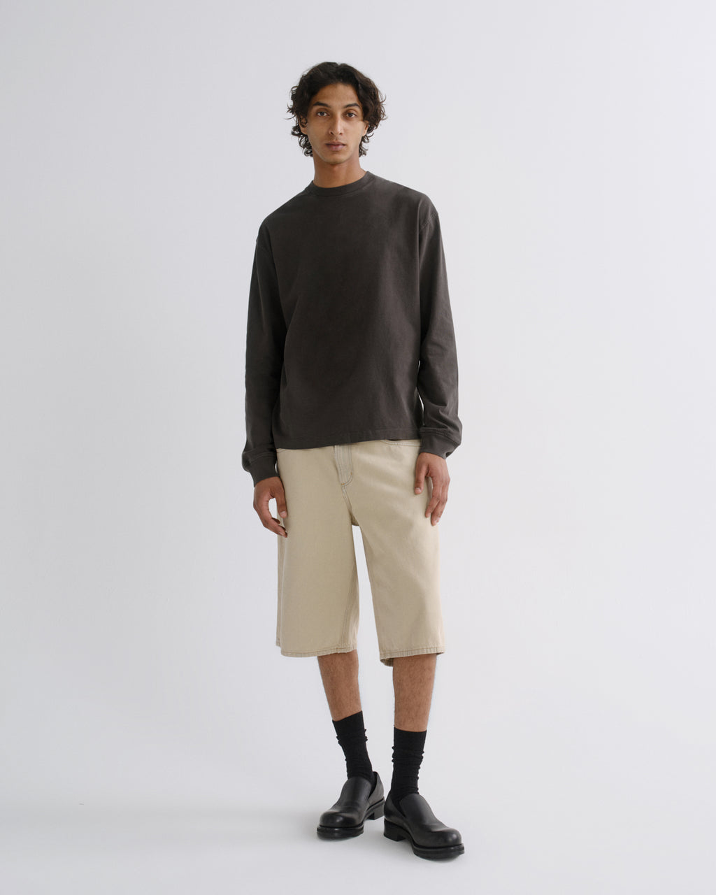 A model stands against a light gray studio background wearing a dark charcoal long-sleeve crewneck sweatshirt, beige knee-length shorts, black socks, and black leather shoes.