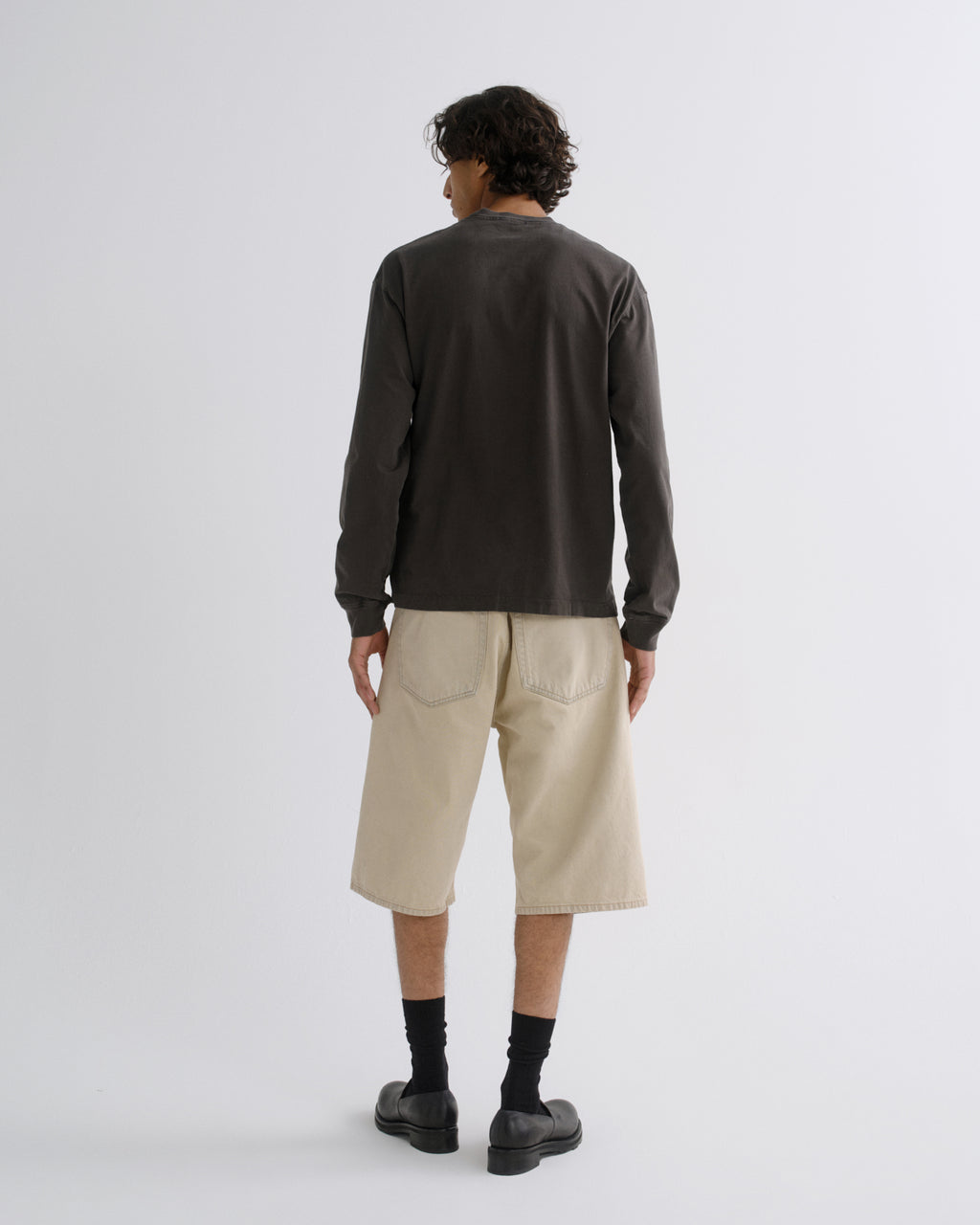 A man is shown from the back wearing a dark charcoal long-sleeve t-shirt, beige cropped shorts, black socks, and black shoes, standing against a light gray studio background.