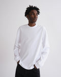 A man wearing a plain white long-sleeve crewneck shirt and black pants, standing against a light gray studio background.