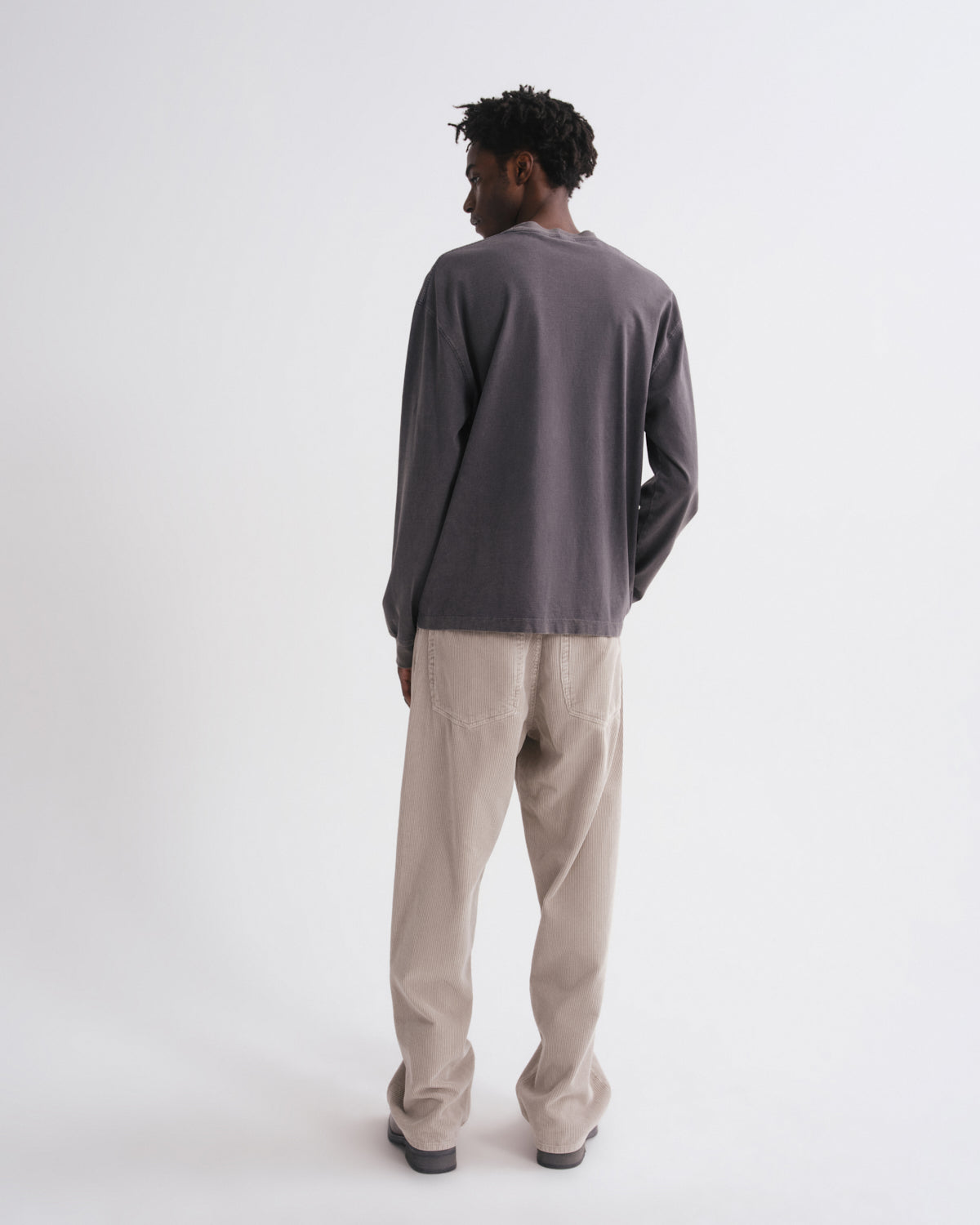 Back view of man wearing a dark grey long-sleeve shirt and beige pants on a grey background