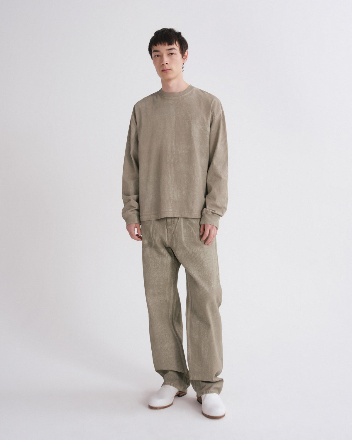 A model stands against a light gray studio background wearing a matching two-piece lounge set in a muted taupe-beige corduroy fabric, consisting of a long-sleeve crewneck sweatshirt and wide-leg pants, paired with white shoes.