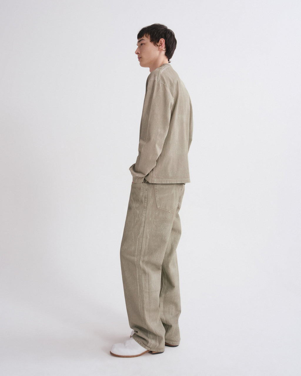 A man stands in profile wearing a light beige corduroy jacket and matching wide-leg trousers, paired with white slip-on shoes, against a plain light gray studio background.