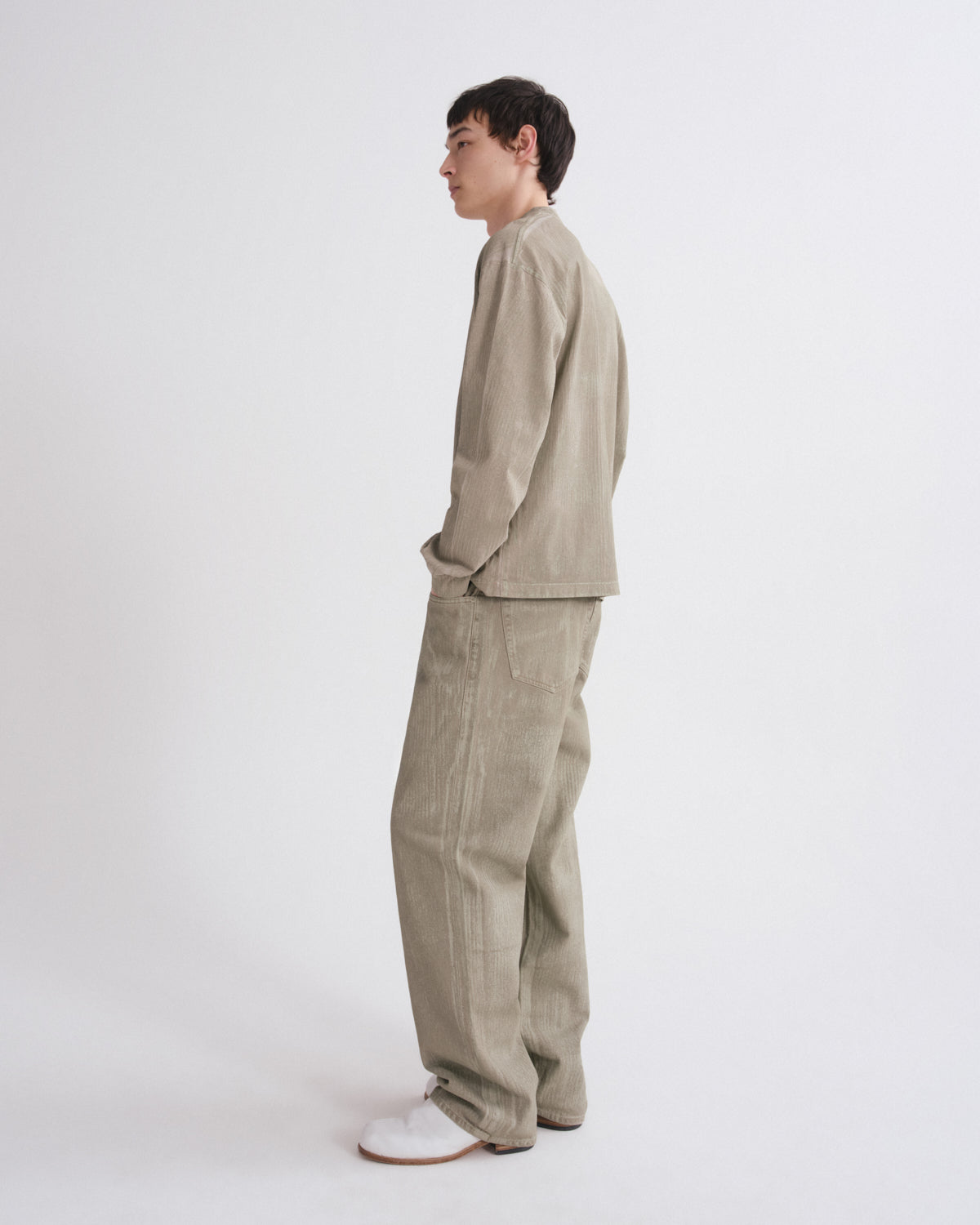 A man stands in profile wearing a light beige corduroy jacket and matching wide-leg trousers, paired with white slip-on shoes, against a plain light gray studio background.