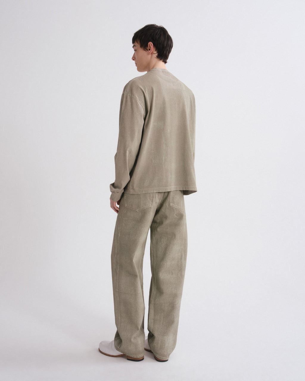 A model shown from the back wearing a matching light beige linen two-piece set consisting of a long-sleeve top and wide-leg trousers, photographed against a seamless light gray studio background.