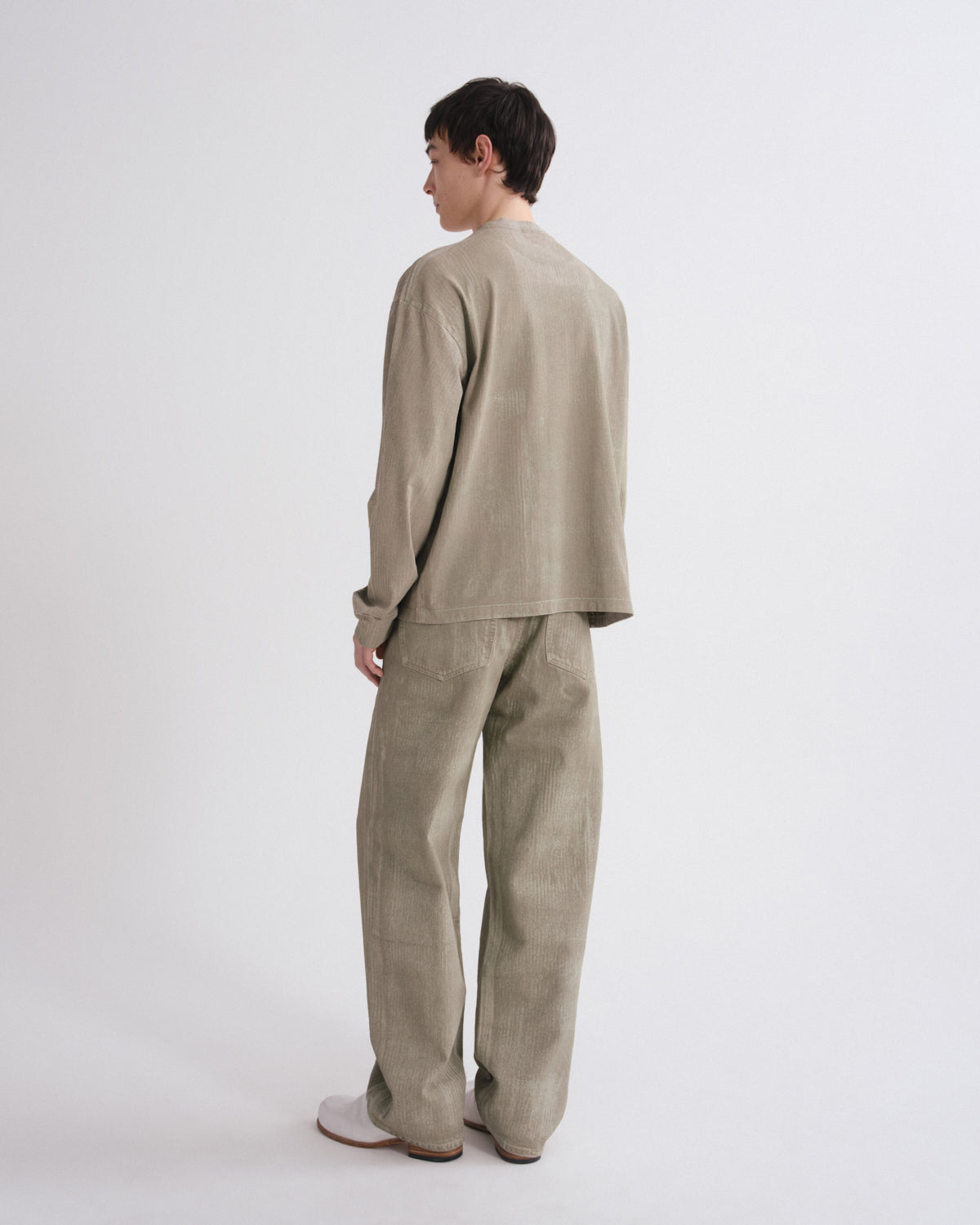 A model shown from the back wearing a matching light beige linen two-piece set consisting of a long-sleeve top and wide-leg trousers, photographed against a seamless light gray studio background.