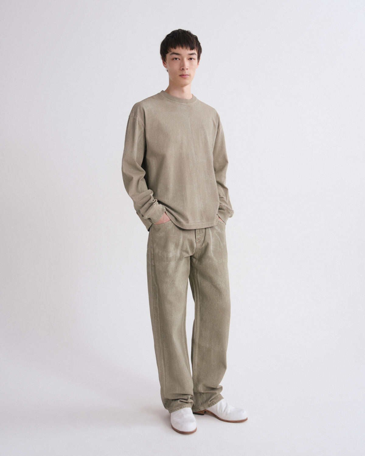 A man modeling a matching beige corduroy long-sleeve shirt and trousers set, paired with white slip-on shoes, against a plain light gray studio background.