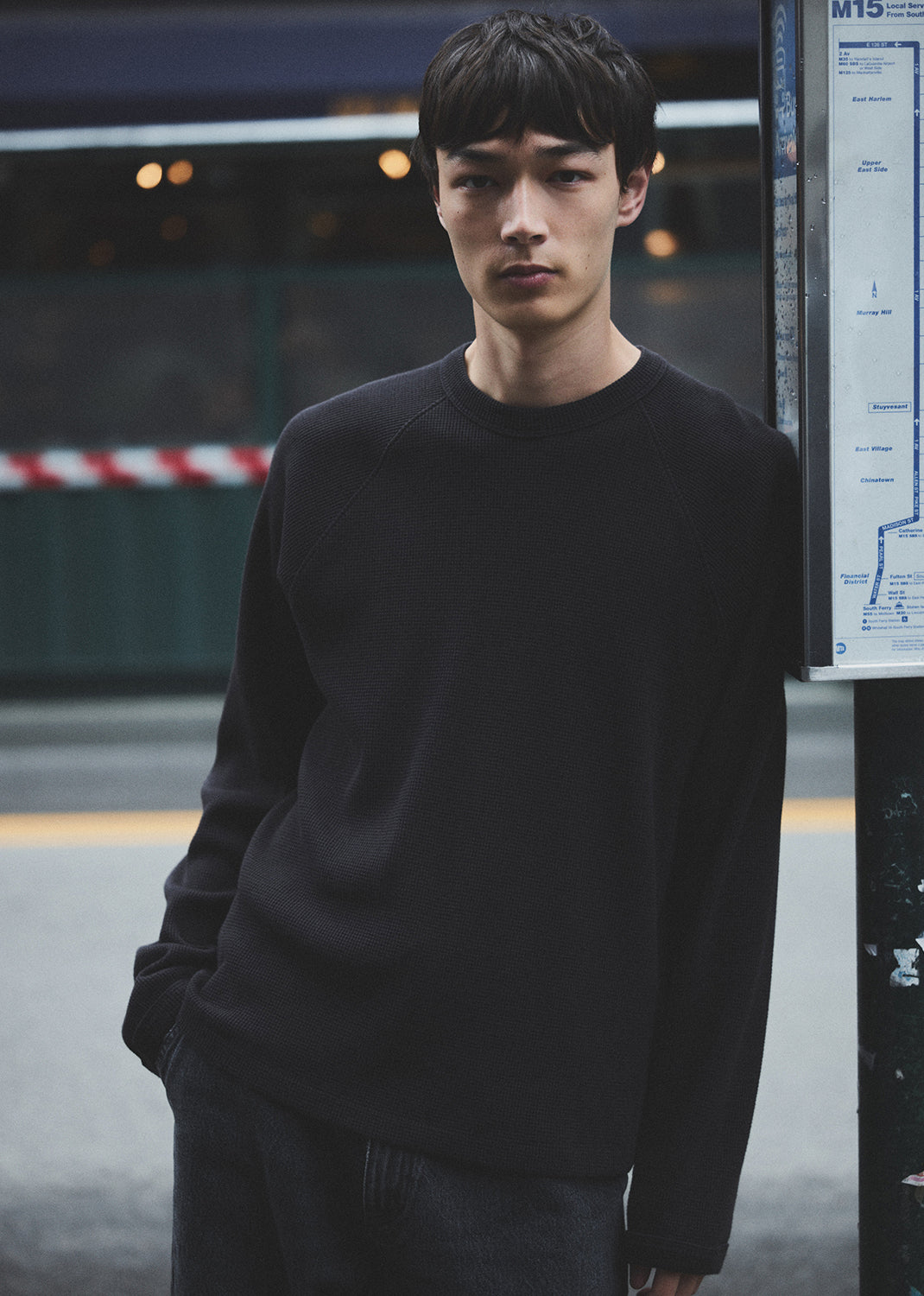 Man wearing a black sweater standing in an urban setting with a bus stop sign in the background.