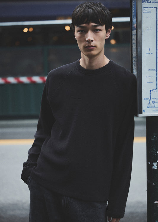 Man wearing a black sweater standing in an urban setting with a bus stop sign in the background.