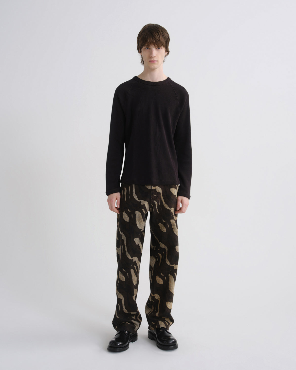 A model stands against a light gray studio background wearing a black long-sleeve crewneck sweater, loose-fit camouflage pants in black and tan, and black leather shoes.