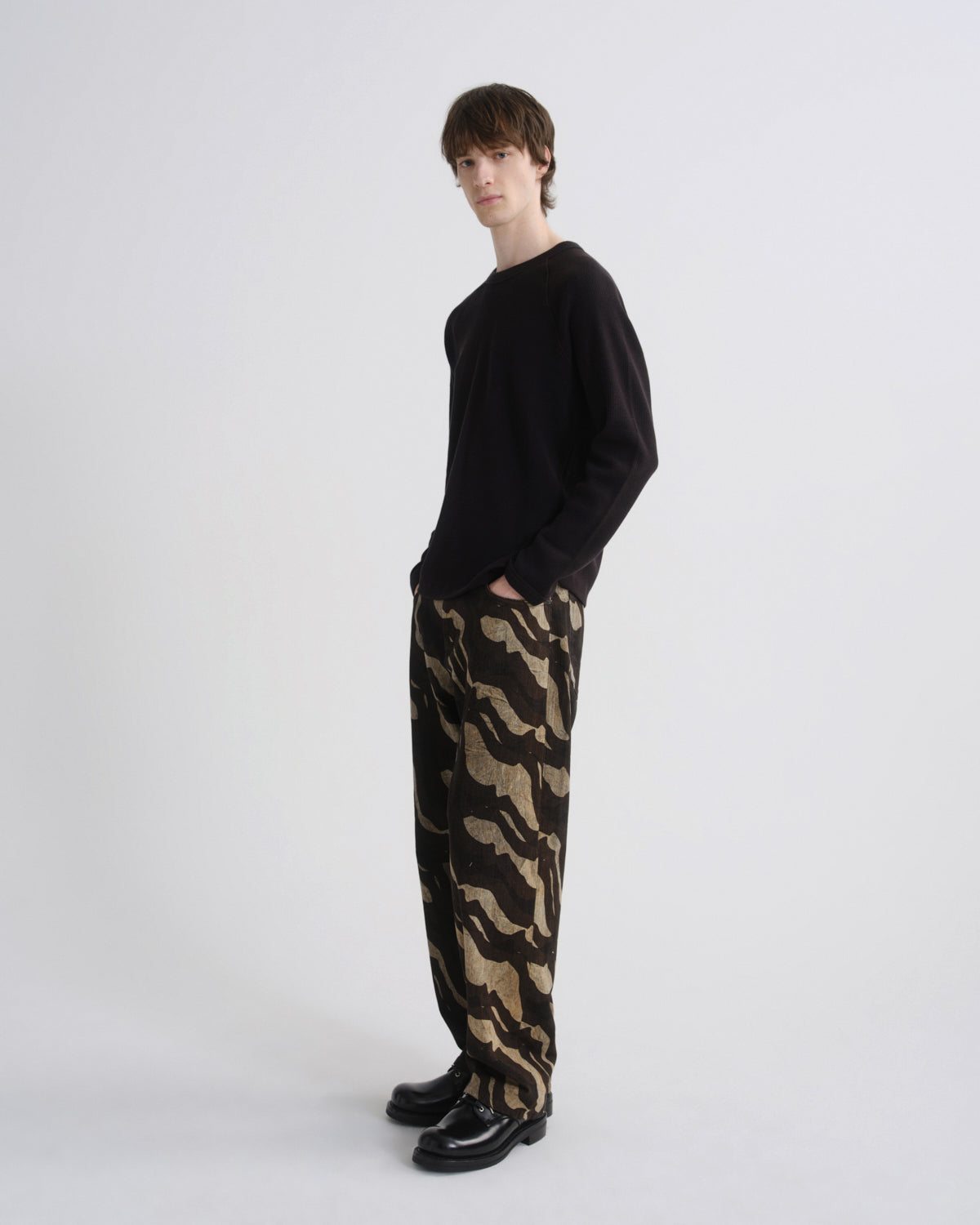 A model stands in profile wearing a black long-sleeve knit top and loose black trousers with a beige abstract wavy pattern, paired with black leather shoes, against a plain light gray studio background.