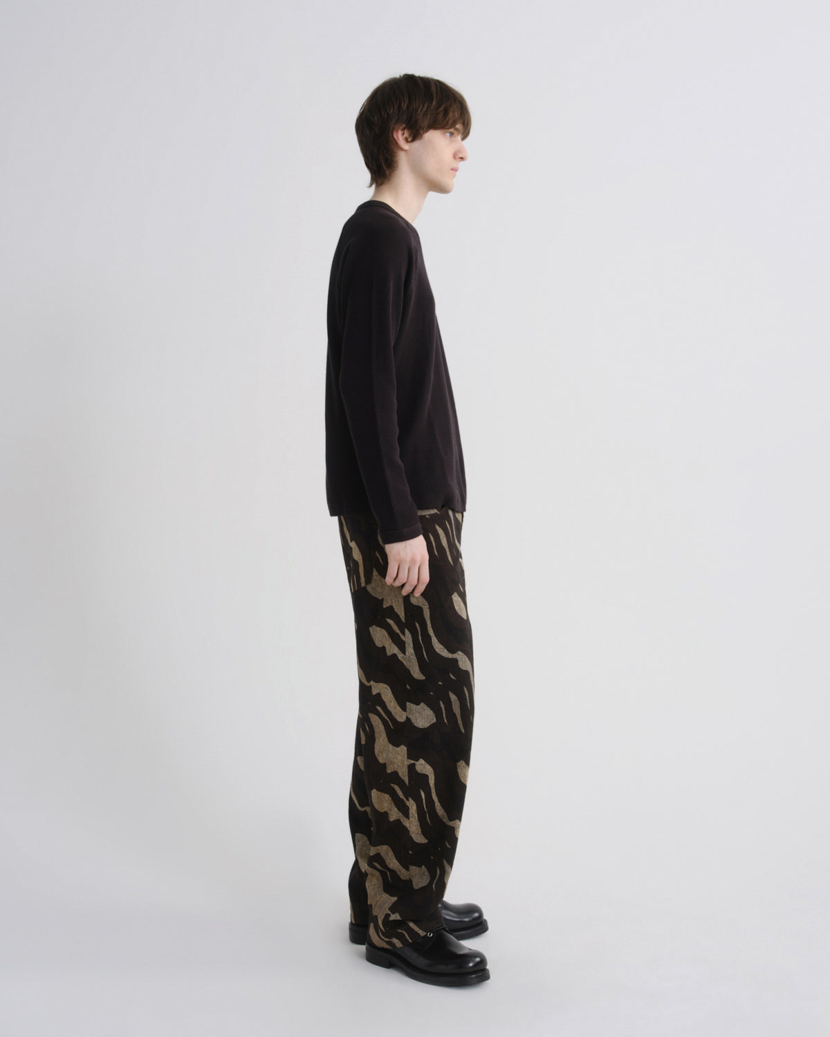 A model standing in profile wearing a black long-sleeve knit top and wide-leg trousers with a black and tan camouflage pattern, paired with black leather shoes, against a light gray studio background.