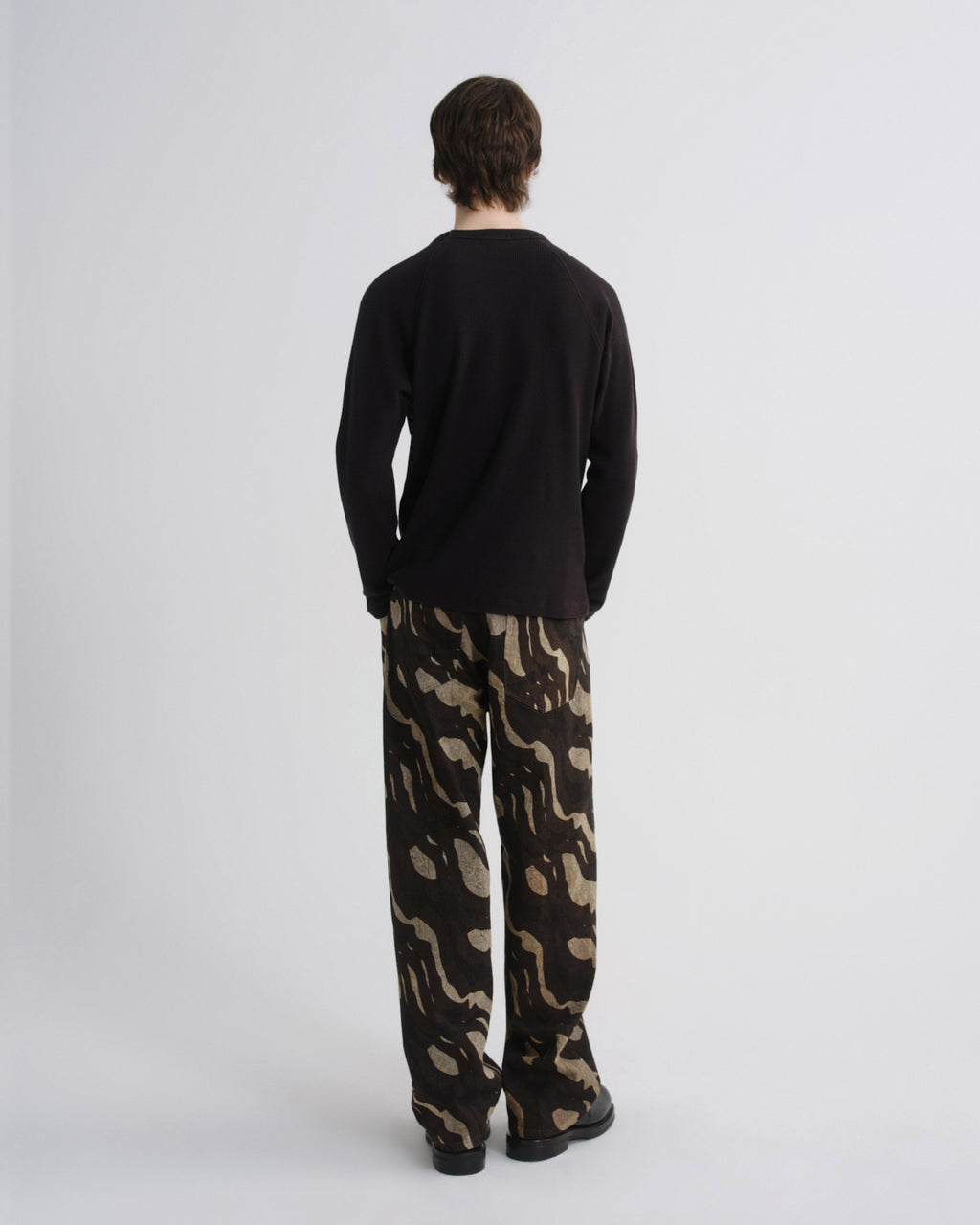 A man viewed from behind wearing a black long-sleeve shirt and loose-fit trousers with a beige and black abstract camouflage pattern, standing against a light gray studio background.