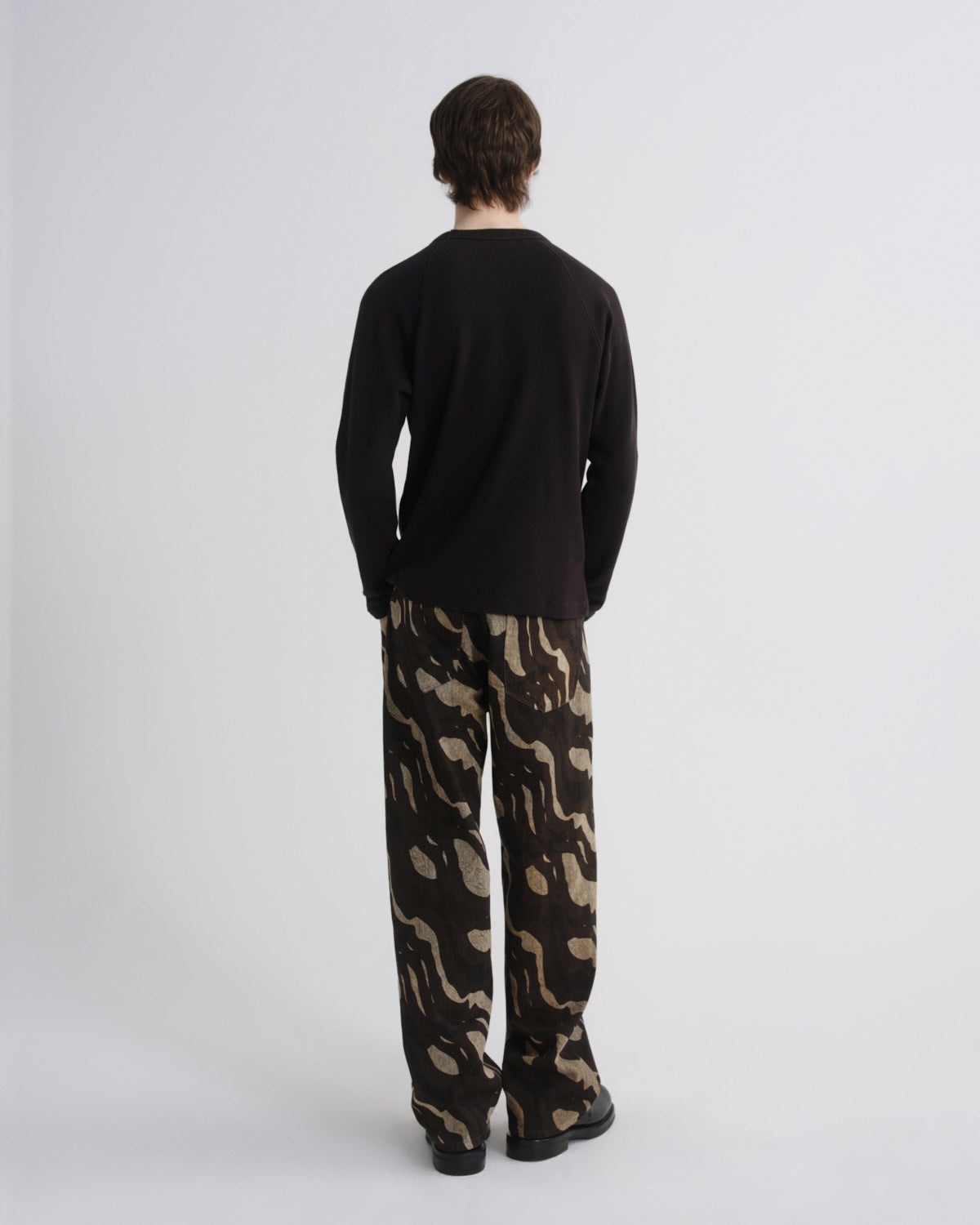 A man viewed from behind wearing a black long-sleeve shirt and loose-fit trousers with a beige and black abstract camouflage pattern, standing against a light gray studio background.