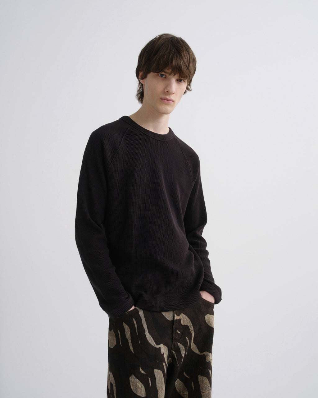 A young man modeling a black long-sleeve crewneck sweatshirt and black pants with a beige abstract pattern, standing against a light gray studio background.