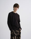 A young man modeling a black long-sleeve crewneck sweatshirt and black pants with a beige abstract pattern, standing against a light gray studio background.