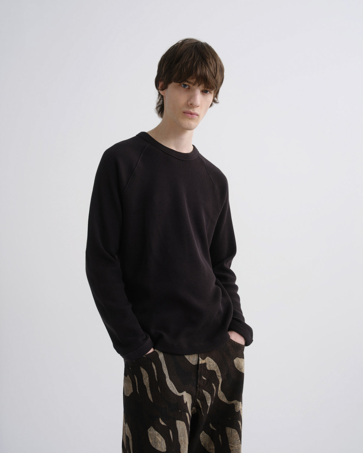 A young man modeling a black long-sleeve crewneck sweatshirt and black pants with a beige abstract pattern, standing against a light gray studio background.
