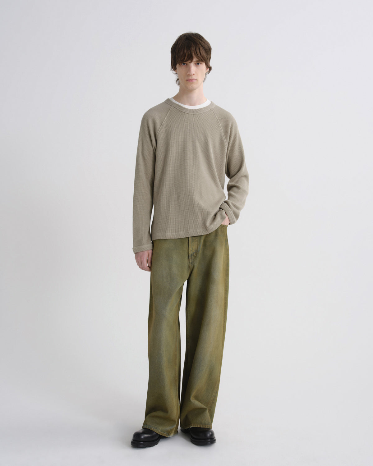 A model stands against a light gray studio background wearing a beige long-sleeve crewneck sweater over a white undershirt, paired with olive green wide-leg trousers and black shoes.