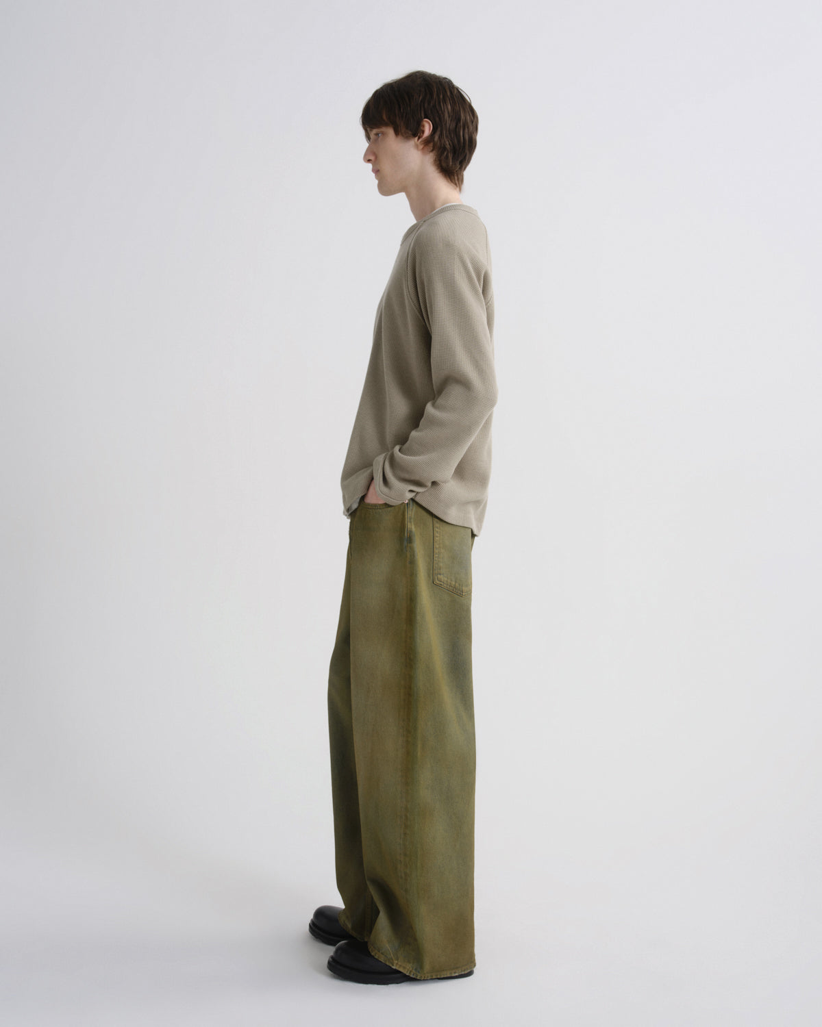 A model stands in profile wearing a light beige long-sleeve knit sweater and wide-leg olive green trousers against a plain white studio background.
