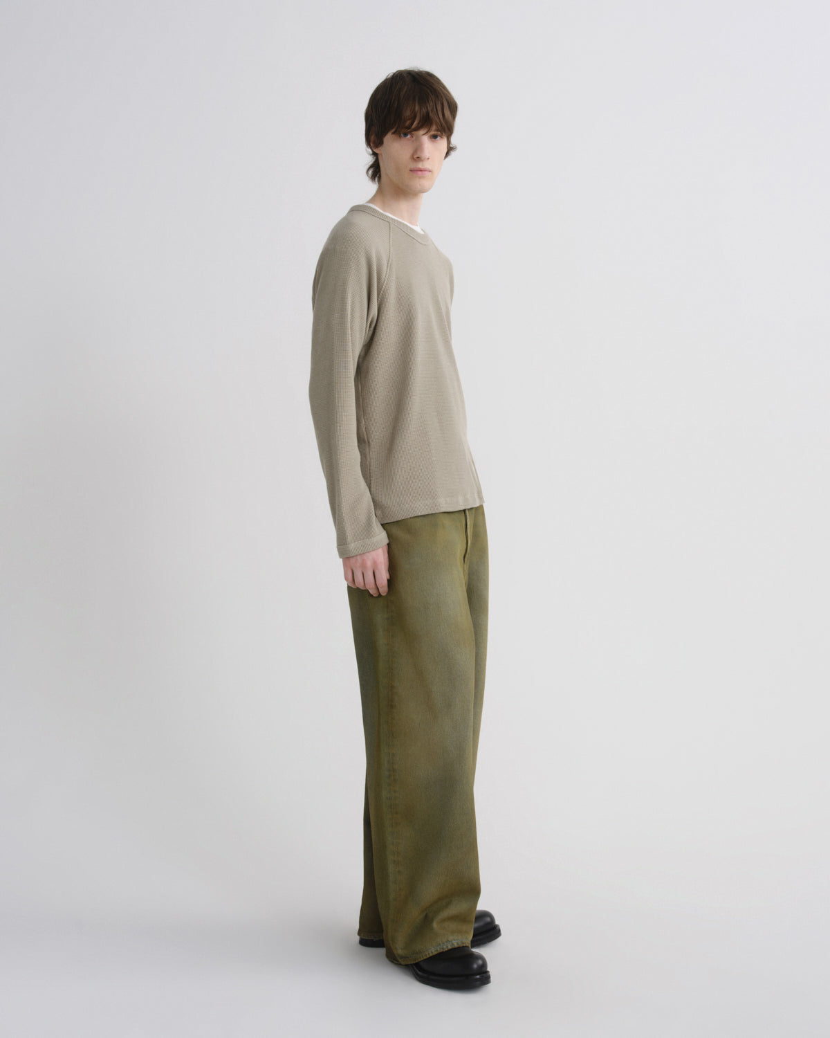 A model stands in profile wearing a light beige long-sleeve knit sweater and olive green wide-leg trousers against a plain light gray studio background.
