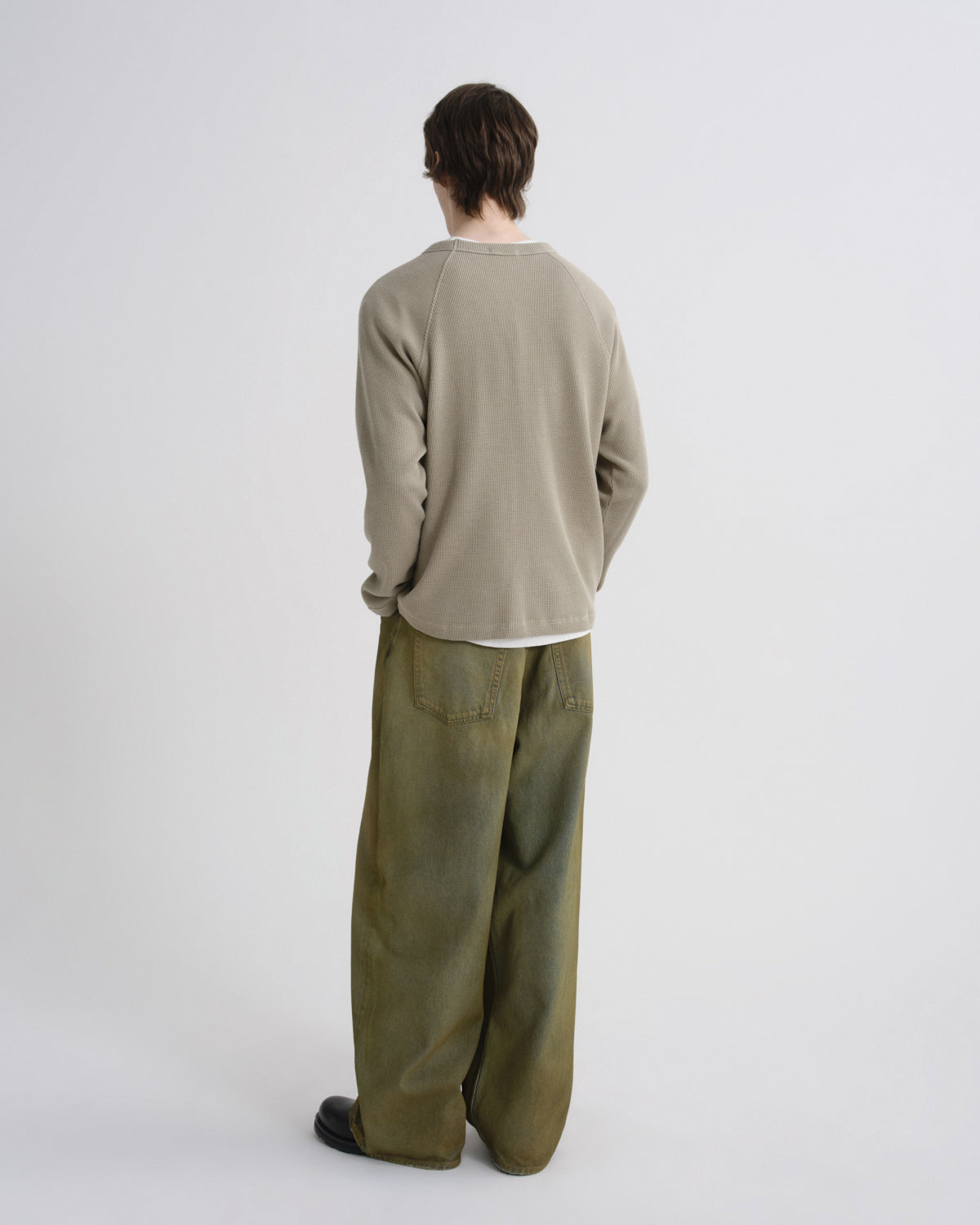 A model shown from the back wearing a light beige long-sleeve knit top and wide-leg olive green trousers, standing against a plain light gray studio background.