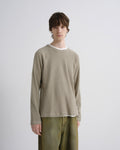 A male model wearing a light beige long-sleeve crewneck sweatshirt over a white t-shirt and olive green pants, standing against a light gray studio background.