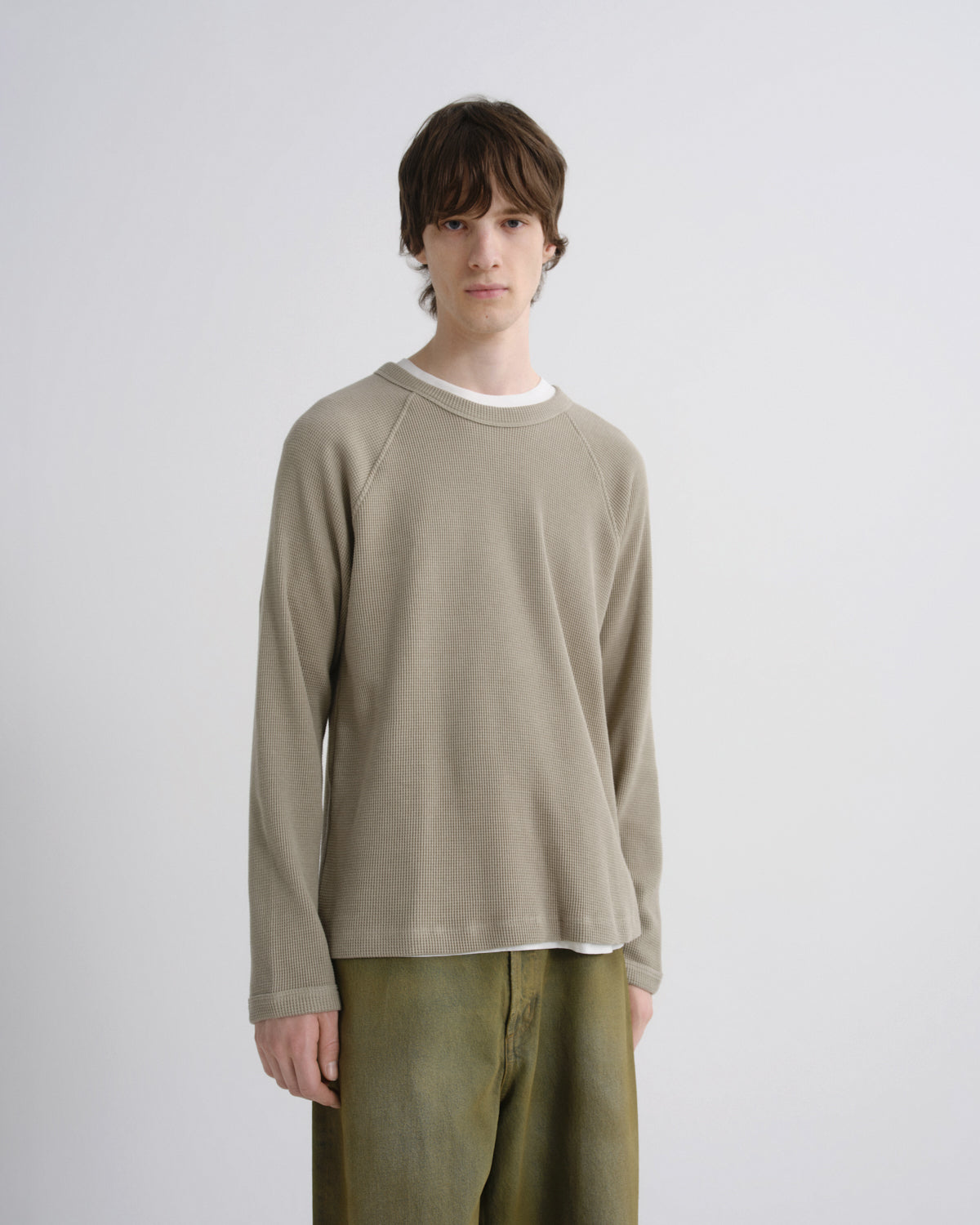 A male model wearing a light beige long-sleeve crewneck sweatshirt over a white t-shirt and olive green pants, standing against a light gray studio background.