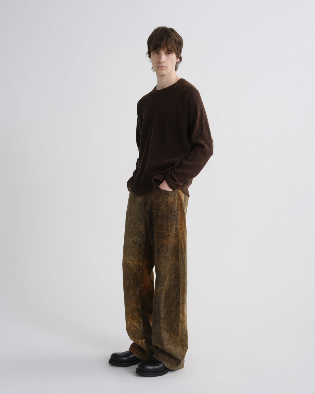 A model stands against a light gray studio background wearing a dark brown long-sleeve knit top, wide-leg brown pants with a subtle mottled pattern, and black shoes.
