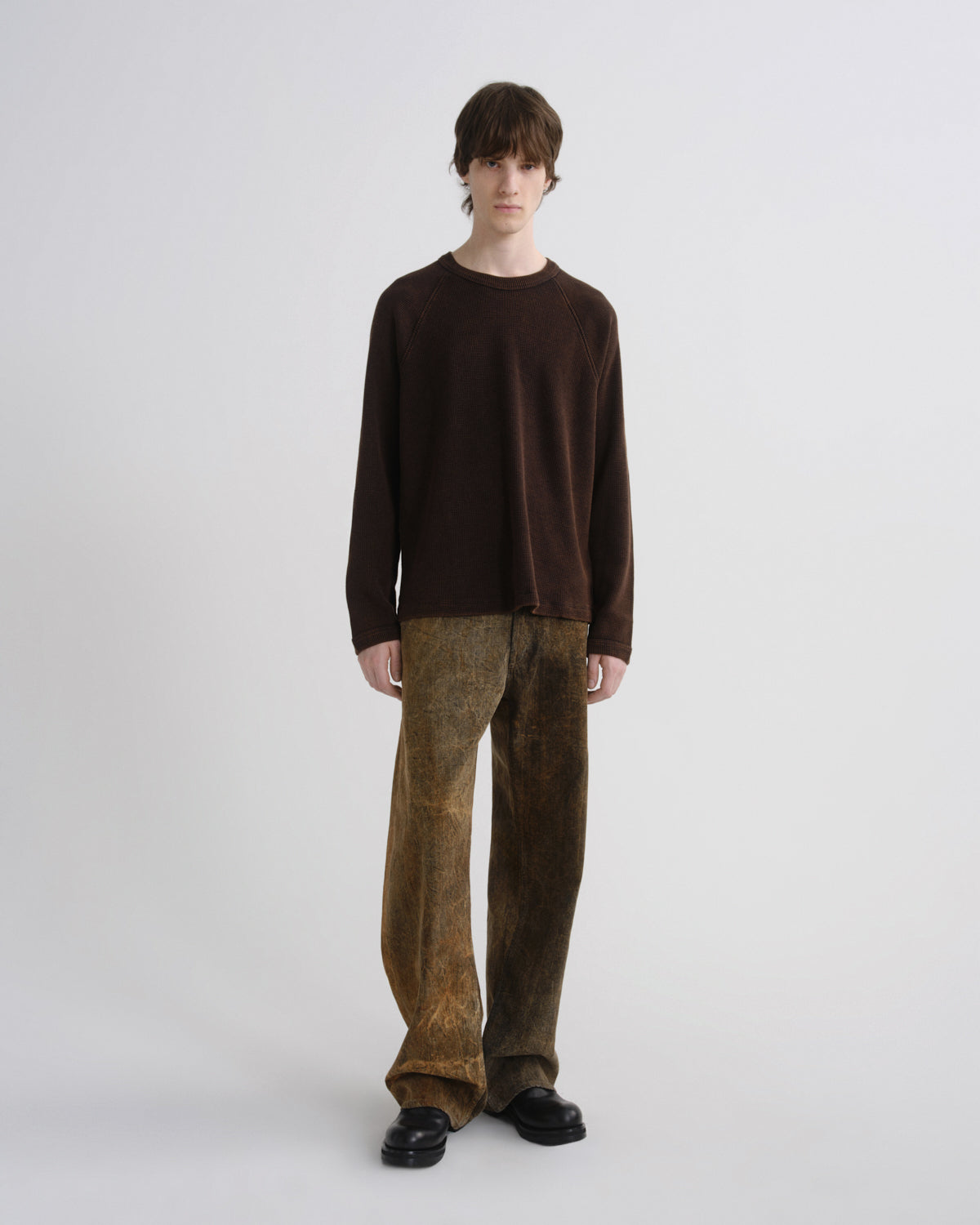 A model stands against a light gray studio background wearing a dark brown long-sleeve knit top, brown textured wide-leg trousers, and black shoes.
