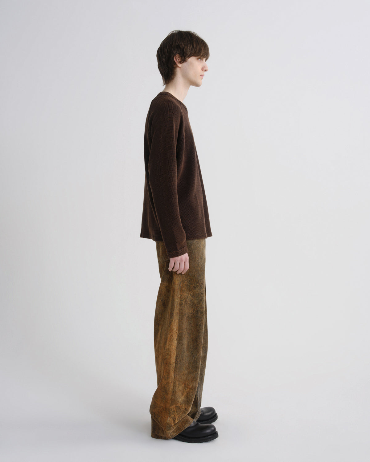 A model shown in profile wearing a dark brown long-sleeve knit top and wide-leg brown trousers, standing against a plain light gray studio background.