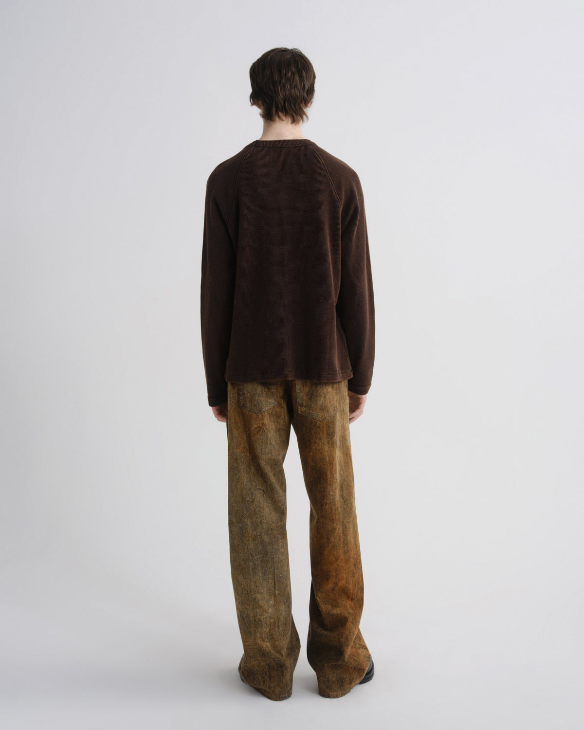 A model shown from the back wearing a dark brown long-sleeve knit top and wide-leg brown pants with a mottled, distressed texture, set against a plain light gray studio background.