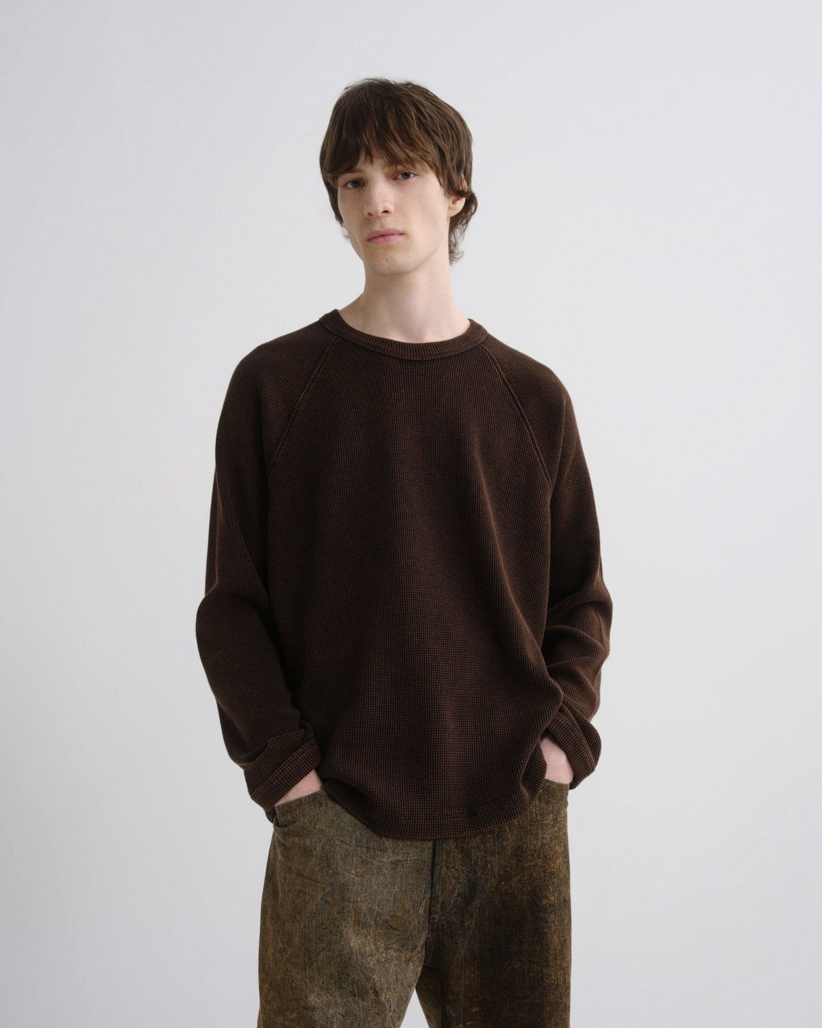 A man wearing a dark brown long-sleeve crewneck sweatshirt and brown patterned trousers, standing against a light gray studio background.